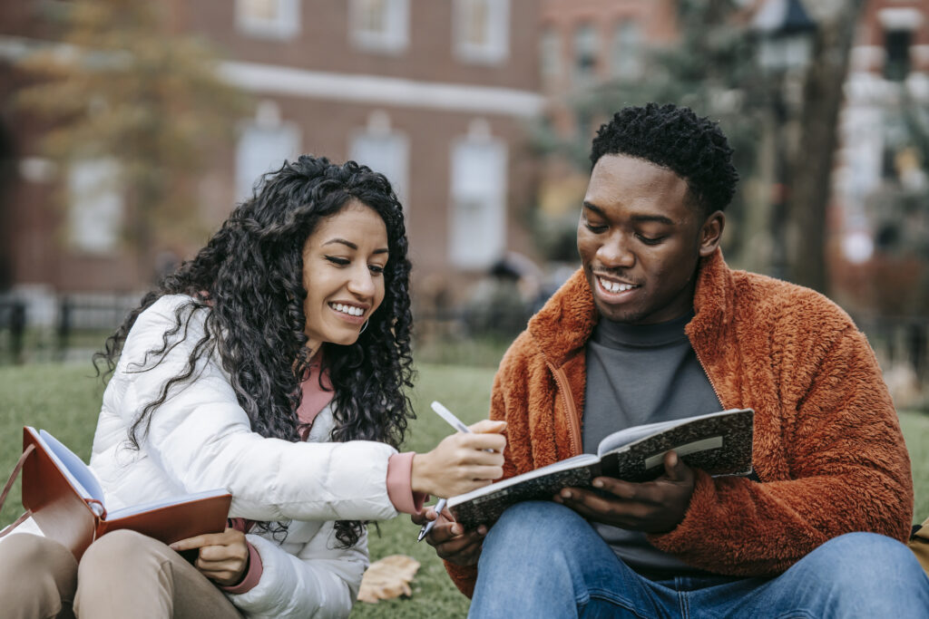 Man and woman holding notebooks while sitting on grass after moving into college